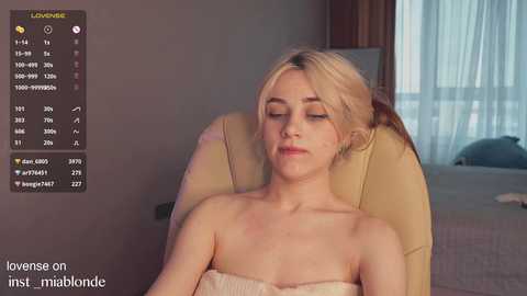 Media: Video of a blonde woman in a beige chair, wearing a strapless, light-colored top, with a background of a bed and window with sheer curtains. Text overlay includes stats about her body measurements and age.