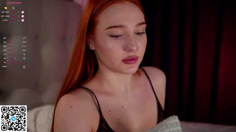 Video of a fair-skinned woman with long red hair, wearing a black bra, lying on a bed. She has closed eyes, and the background is dark and blurry.