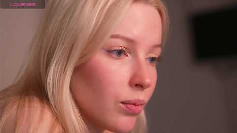 Video of a young, fair-skinned blonde woman with light blue eyes, wearing minimal makeup, looking introspective. Background is blurred, featuring a beige wall and a dark TV screen.