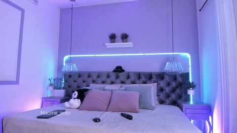 Media: Video of a chic, purple-themed bedroom with a tufted grey headboard, LED strip lights, two nightstands with potted plants, and a plush bed with pink pillows.