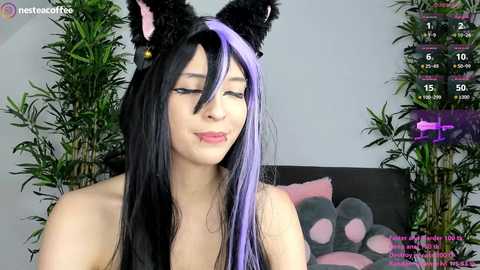 Video of an Asian woman with long black and purple hair, wearing black cat ears, smiling in a playful manner. Background includes a potted plant and a black cushion with a pink paw print.