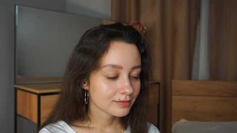 Media: Video of a young woman with long brown hair, wearing a white shirt and hoop earrings, in a bedroom with wooden furniture and beige curtains.