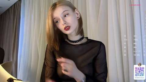 Video of a young Caucasian woman with light skin and shoulder-length blonde hair, wearing a sheer black top and black choker, looking pensive against beige curtains.