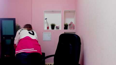 A video of a pink-walled office with a person in a red and white sweater, seated at a black desk, facing away. Two small white shelves with plants and a pink vase are visible.