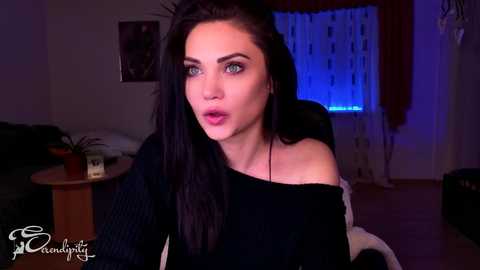 A video of a woman with long black hair, green eyes, and light skin, wearing a black off-shoulder sweater, in a dimly lit room with a blue-lit background.