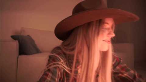 Video of a fair-skinned woman with long, straight blonde hair, wearing a brown hat and plaid shirt, smiling, sitting on a beige couch with a gray cushion. Warm, reddish lighting.