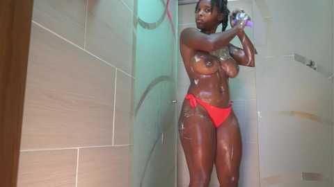 Media: Video of a dark-skinned woman with long hair, medium-sized breasts, and a curvy physique, wearing red bikini bottoms, showering in a tiled bathroom.