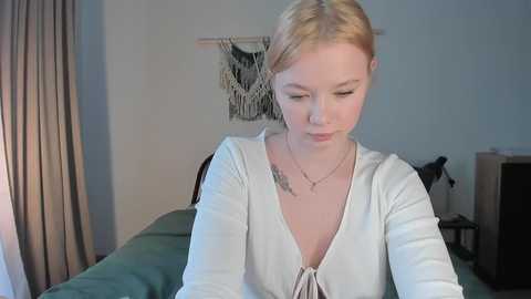 Media: Video of a fair-skinned, blonde-haired woman in a white V-neck sweater, sitting on a green bed, with a woven wall hanging and beige curtains in a dimly lit room.