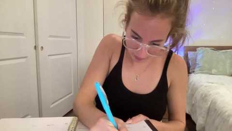 Media: Video of a blonde woman with glasses, wearing a black tank top, writing with a blue pen at a desk in a cozy bedroom with white doors, a bed, and purple lighting.