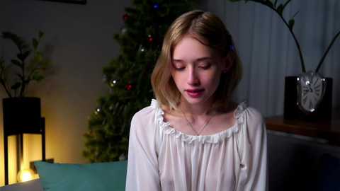 A video of a young woman with fair skin and shoulder-length blonde hair, wearing a sheer white blouse, seated indoors by a lit Christmas tree, with a candle-lit lantern and decorative plant in the background.