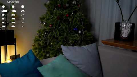 A video of a cozy living room with a lit Christmas tree adorned with ornaments and a wooden clock on a shelf, surrounded by blue and teal pillows on a sofa, illuminated by ambient lighting.