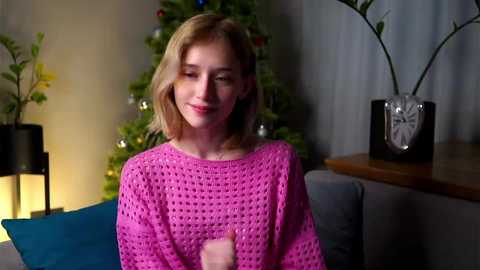 Video of a smiling young blonde woman with fair skin in a pink knitted sweater, standing in a cozy living room with a lit Christmas tree, potted plants, and a wooden side table.