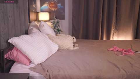 Video of a cozy bedroom with a beige bed, white pillows, a pink dress, and a white lamp on a nightstand, framed by beige curtains.