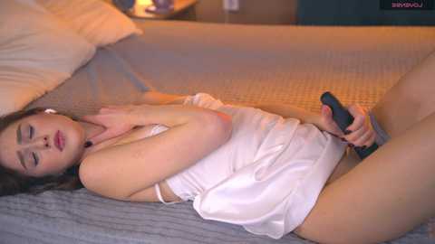 A video of a fair-skinned woman with brown hair, wearing a white camisole and panties, lying on a bed, masturbating with a black vibrator.