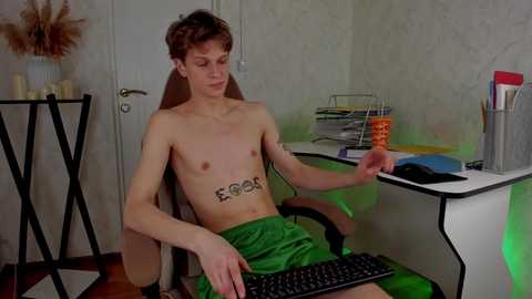 Media: Video of a shirtless, lean, white male with short brown hair, wearing green shorts, seated at a desk with a computer and office supplies in a dimly lit room with floral wallpaper.