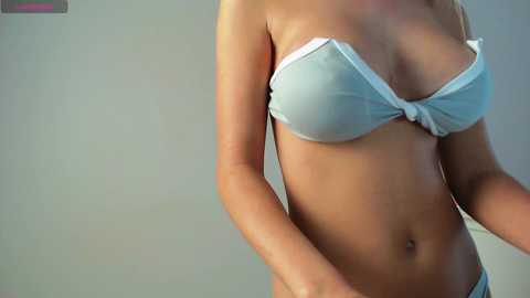 Media: Video of a light-skinned woman wearing a light blue, strapless bra that accentuates her medium-sized breasts. Her smooth, toned abdomen is visible, and she stands against a plain, light gray wall.