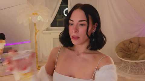 Media: Video of a young woman with straight, shoulder-length black hair, wearing a white off-shoulder top, sitting in a cozy, dimly-lit room with beige curtains, a white chair, and a yellow lamp.