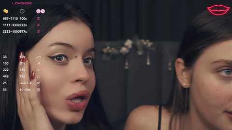 Video of two young women with fair skin, one with dark hair, wearing makeup, and the other with light hair, both smiling, in a dimly-lit room.