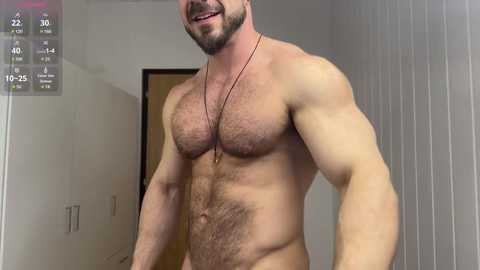 Media: Video of a muscular, bearded man with a hairy chest and light skin, standing indoors in a hallway. He wears a necklace and has a slight smile.