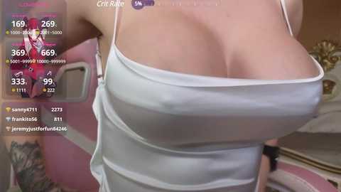 Video of a woman with light skin, wearing a white, low-cut tank top that reveals her large, cleavage-enhancing breasts. Background shows a pink room with a bed and a smartphone screen displaying chat messages.