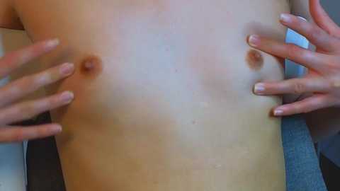 Media: Video of a fair-skinned person with small, round breasts, hands cupping the breasts from either side. Background is blurred, showing a blue fabric surface.