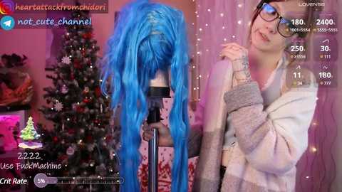 Media: A video of a young woman with blue hair and glasses, wearing a gray sweater, standing beside a Christmas tree in a cozy, decorated room.