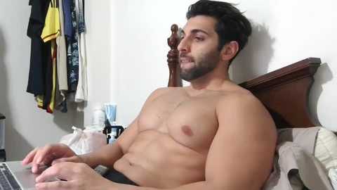 Media: Video of a shirtless, muscular Middle Eastern man with dark hair and beard, sitting on a bed, typing on a laptop, surrounded by clothes on a wooden headboard.