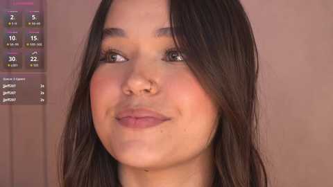 Video of a young woman with long, straight, dark brown hair, light skin, and full lips, wearing minimal makeup. She has a nose ring. Background shows a blurred beige wall and a digital screen displaying weather data.