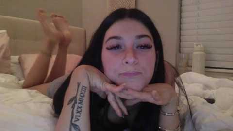 Media: A video of a young woman with long dark hair, lying on a bed, wearing minimal makeup, showcasing tattoos on her arms. The background shows a beige headboard and white blinds.