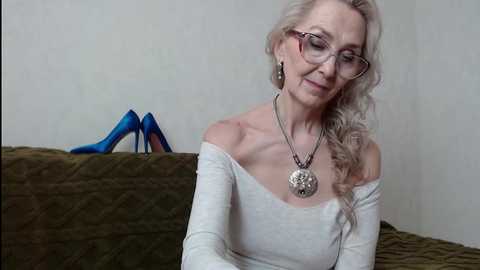 Media: Video of a blonde, middle-aged woman with glasses and long, wavy hair, wearing a white off-shoulder top and a silver pendant necklace. She sits on a brown quilted couch with blue high heels and a gray wall background.