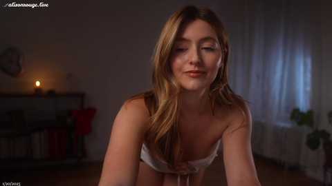 Media: A video of a young, light-skinned woman with long, wavy blonde hair and medium-sized breasts, wearing a white strapless bra, smiling softly in a dimly lit room with a lit candle, a clock, and a potted plant in the background.