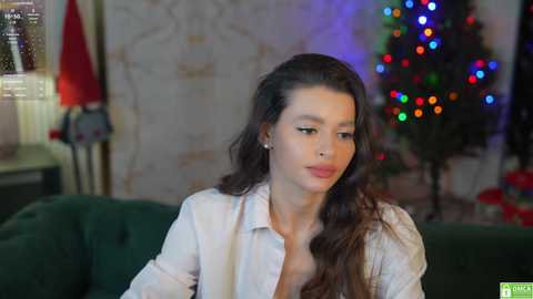 Media: A video of a young woman with long, dark hair, wearing a white shirt, sitting on a green couch, surrounded by festive lights and a decorated Christmas tree in a cozy living room.