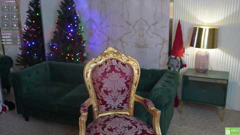 Media: Video of a lavish living room with a gold, red-patterned chair facing a green velvet sofa, Christmas trees with multicolored lights, and a modern lamp on a white wainscoted wall.