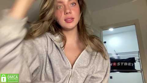 Media: Video of a young Caucasian woman with long, wavy blonde hair, fair skin, and green eyes, wearing a grey robe, standing in a modern, brightly-lit bathroom.