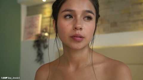 Video of a young woman with light skin, dark hair in a bun, and bare shoulders, standing indoors against a blurred background.