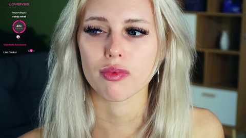 Media: Video of a young, fair-skinned woman with platinum blonde hair, blue eyes, and full lips. She has a neutral expression. Background includes a dark couch and wooden shelves with blue items.