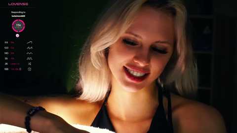 A video of a smiling, light-skinned blonde woman with shoulder-length hair, wearing a black halter top, indoors with dim lighting, on a video call.