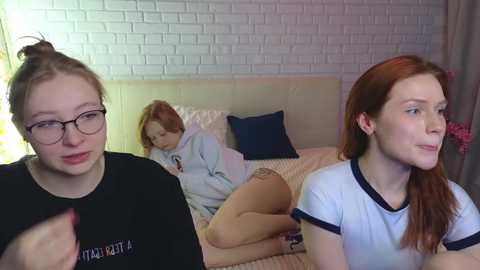 Video of three young women in a cozy bedroom; one blonde in glasses, one redhead, and one brunette, all with fair skin, wearing casual attire.