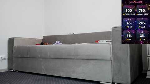 Media: Video of a grey, minimalist sofa with a single pillow, against a white wall in a sparsely decorated room. Right corner features a digital leaderboard with various scores.