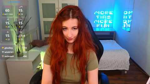 Media: Video of a fair-skinned woman with long, wavy red hair, wearing a green t-shirt, seated in a gaming chair in a modern bedroom with blue neon lights and a bed.