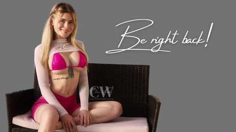 Video of a smiling, blonde woman with fair skin and a medium build, wearing a pink bikini top and shorts, sitting on a wicker chair with a gray background. Text reads \"Be right back!\