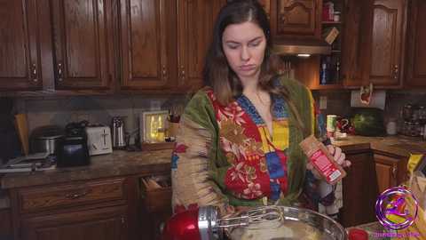 Media: Video of a woman in a colorful, patchwork robe mixing cake batter in a wooden kitchen. Wooden cabinets, appliances, and a watermelon are in the background.