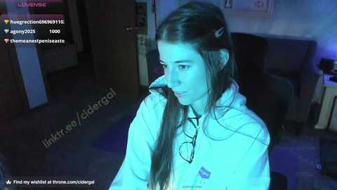 Media: A young woman with long brown hair, wearing a light hoodie, gazes intently at a video game controller in a dimly lit room.