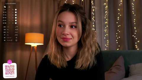 Media: Video of a young Caucasian woman with shoulder-length wavy blonde hair, smiling, wearing a black top. Background features a teal couch, beige curtains, a warm-toned lamp, and a digital overlay with \"LOUNGE\" text.