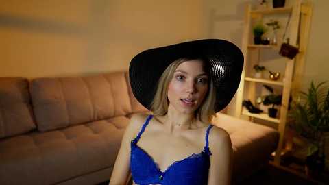 Media: Video of a blonde woman in a blue lace bra and a large black cowboy hat, standing in a cozy living room with beige sofa and wooden shelving unit.