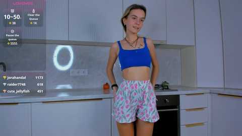 Media: Video of a young Caucasian woman with light skin and blonde hair, wearing a blue crop top and floral shorts, standing in a modern white kitchen.