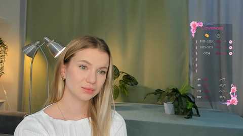 Media: Video of a young Caucasian woman with straight blonde hair, wearing a white top, standing in a modern room with a green curtain, plant, and digital clock display.