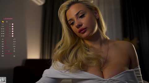 Video of a blonde woman with long hair, wearing an off-shoulder white shirt, in a dimly lit room, with a TV showing a weather forecast in the background.