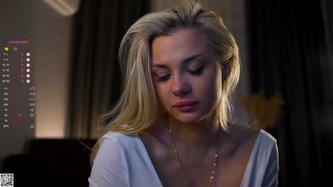 Video of a blonde woman with long hair, wearing a white off-shoulder top, looking contemplative, in a dimly lit room with dark curtains and a digital clock displaying 3:26 AM.