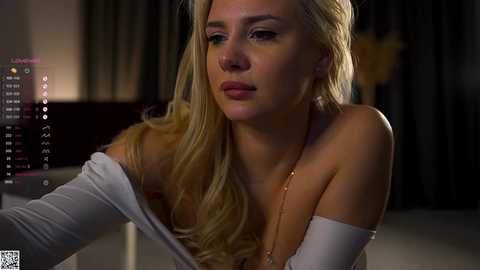 Video of a blonde woman with fair skin, wearing a white off-shoulder top, looking contemplative. Background features a dimly lit room with dark curtains and a glowing screen displaying text.